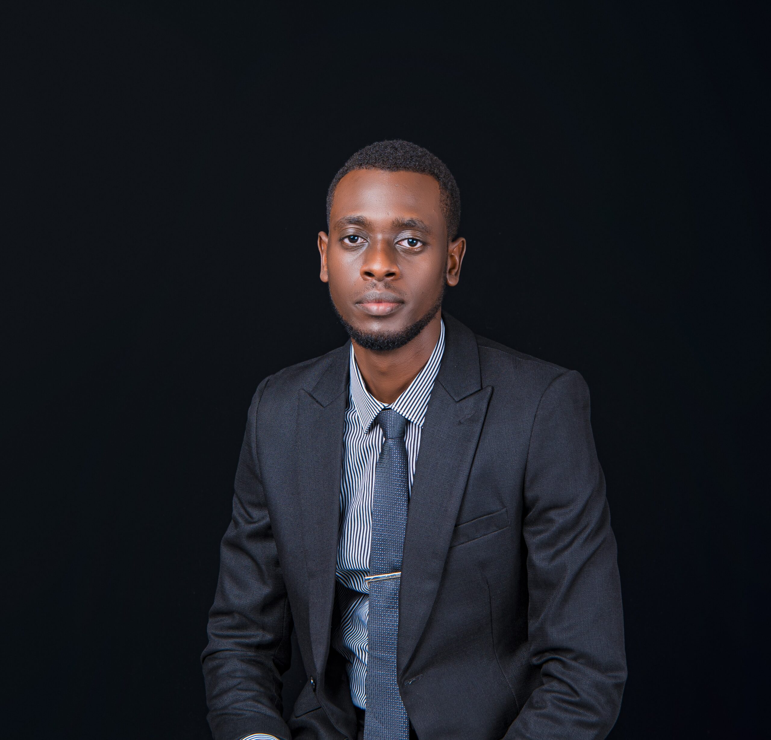 Kevin Ntwari poses in a black suit, grey shirt and tie for portrait photo with a dark background